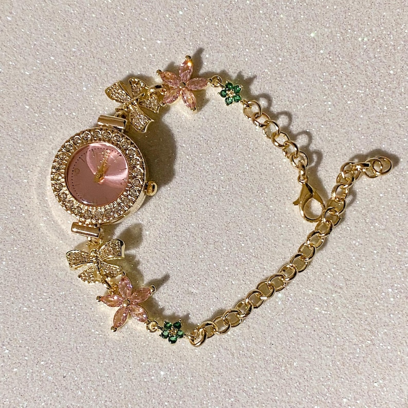 May include: Gold-toned watch bracelet with a pink watch face and a bezel encrusted with small crystals. The bracelet features bow and flower accents with pink and green gemstones. The watch has a chain-link band.