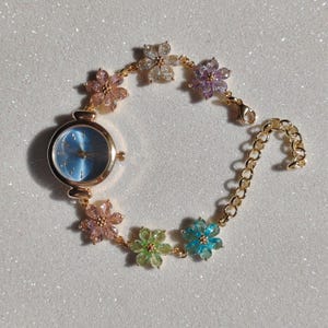May include: A gold-toned wristwatch with a light blue face and a floral bracelet. The bracelet features colorful flower-shaped embellishments in pink, green, blue, purple, and clear. The watch has a classic round face and a delicate chain closure.