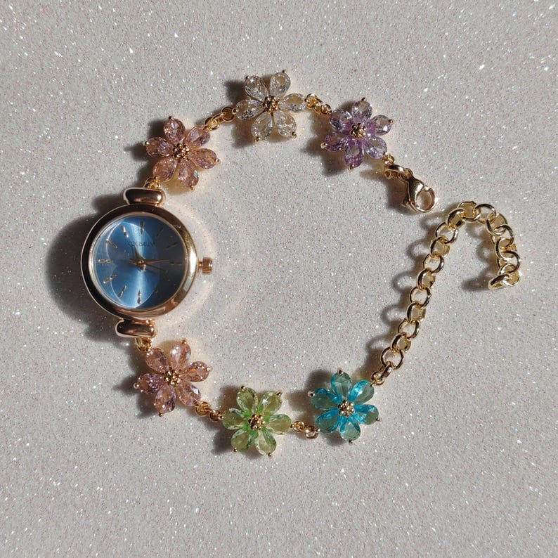 May include: A gold-toned wristwatch with a light blue face and a floral bracelet. The bracelet features colorful flower-shaped embellishments in pink, green, blue, purple, and clear. The watch has a classic round face and a delicate chain closure.