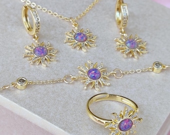 May include: A gold jewelry set consisting of a necklace, a pair of hoop earrings, a bracelet, and a ring. Each piece features a sunburst design with a central round purple opal-like stone surrounded by small clear crystals on a gold-tone metal chain.