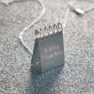 May include: A silver pendant necklace with a notebook-shaped charm. The charm has metal loops at the top and the words "Robbin's Flower Shop" engraved on the front. The necklace chain is also silver. The pendant is a small, detailed design.
