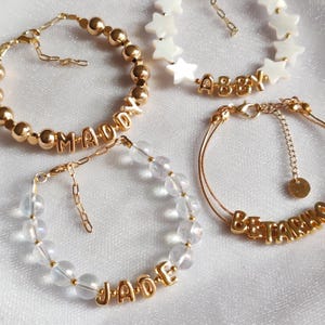 Puede incluir: Cuatro pulseras de oro con diferentes encantos. La primera pulsera tiene cuentas doradas y el nombre "MADDY" escrito en letras doradas. La segunda pulsera tiene cuentas transparentes iridiscentes y el nombre "JADE" escrito en letras doradas. La tercera pulsera tiene una cadena de oro con el nombre "ABBY" escrito en letras doradas. La cuarta pulsera tiene una cadena de oro con el nombre "BETTY" escrito en letras doradas.
