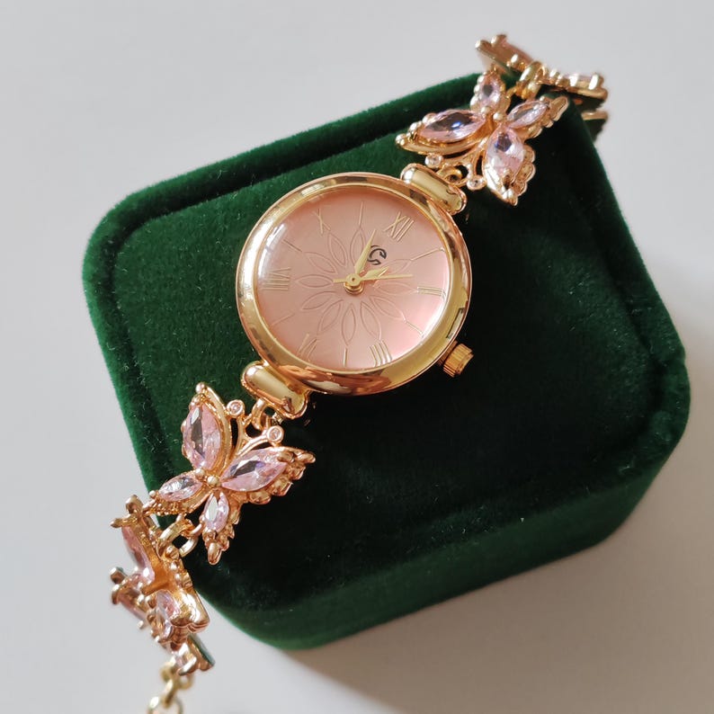 Puede incluir: Un reloj de pulsera dorado con esfera rosa y n&uacute;meros romanos. La correa presenta detalles de mariposas con gemas rosas. El reloj se exhibe sobre una superficie de terciopelo verde oscuro.