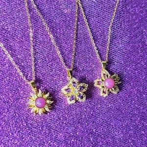 May include: Three gold necklaces with flower-shaped pendants featuring pink and purple gemstones. The necklaces are on a purple sparkly background.