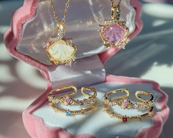Puede incluir: Dos collares de oro con colgantes de concha, uno con una concha blanca y el otro con una concha rosa, y dos anillos de oro con piedras de colores, todo presentado en una caja de terciopelo rosa en forma de concha.