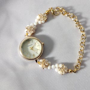 May include: A gold-tone wristwatch with a white face and a gold chain bracelet. The bracelet is adorned with white pearl-like beads and gold flower-shaped accents with clear rhinestones. The watch face has black Roman numeral hour markers and the word "COUSIKUM" printed in black.