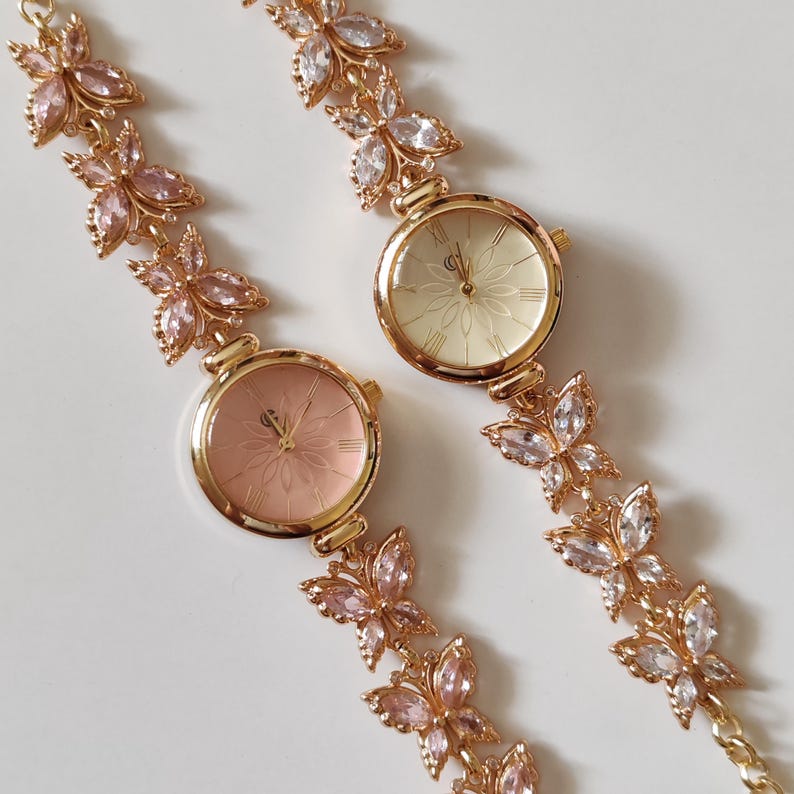 Puede incluir: Dos relojes de pulsera dorados con detalles florales y de mariposas. Las esferas son de color rosa claro y crema, con n&uacute;meros romanos. Las correas presentan adornos en forma de mariposa con cristales rosas y transparentes.