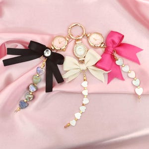 May include: Three gold-tone watches with different colored bows and heart-shaped charms. The watches have white faces and black hands. The bows are black, white, and pink. The charms are made of white and blue iridescent material.