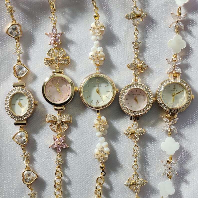 May include: Five gold-tone wristwatches with different decorative charms and crystal accents. The watches have white, pink, and cream-colored faces.
