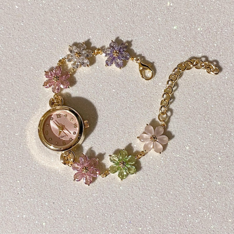 May include: A gold-toned wristwatch with a pink face and a floral bracelet. The bracelet features a series of colorful flower-shaped gemstones in pink, green, purple, and clear. The watch has a quartz movement.