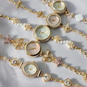 May include: A set of gold-tone wristwatches with delicate floral and heart-shaped charms. Each watch has a white or pink face with black Roman numeral hour markers and a crystal-encrusted bezel.