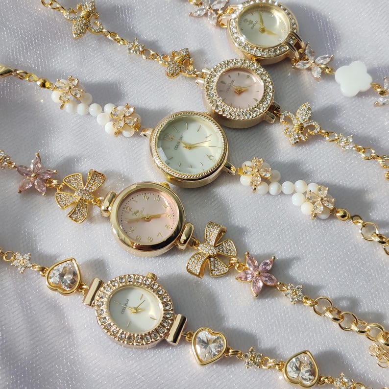 May include: A set of gold-tone wristwatches with delicate floral and heart-shaped charms. Each watch has a white or pink face with black Roman numeral hour markers and a crystal-encrusted bezel.