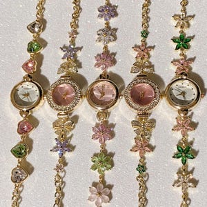May include: Five gold-toned wristwatches with floral and heart-shaped accents. Each watch has a round face with a different colored dial, surrounded by a crystal-studded bezel. The watch bands feature a chain design with colorful flower and heart charms.