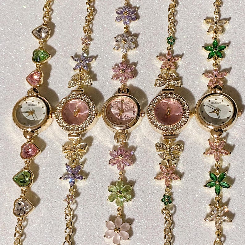 May include: Five gold-toned wristwatches with floral and heart-shaped accents. Each watch has a round face with a different colored dial, surrounded by a crystal-studded bezel. The watch bands feature a chain design with colorful flower and heart charms.