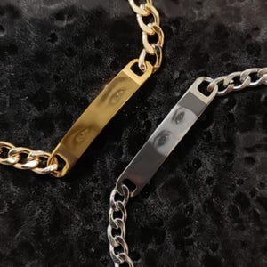 Puede incluir: Dos pulseras de cadena. Una es dorada, la otra plateada. Cada una presenta una placa rectangular con una imagen de ojo. Las pulseras tienen un diseño de eslabones de cadena gruesos y se exhiben sobre una superficie oscura y texturizada.