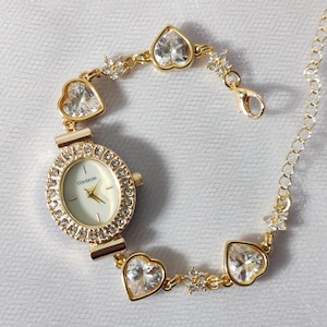 May include: Gold-tone watch with a white face and a crystal-encrusted bezel. The watch is attached to a gold-tone bracelet with heart-shaped charms and crystal accents. The watch face has the word "COUSSEUM" printed on it.