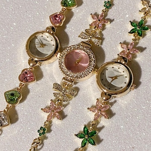 May include: Three gold-toned wristwatches with delicate floral and heart-shaped accents. The watch faces are white and pink, with the brand name "COUSUM" visible on two of them. The bands feature pink, green, and clear gemstones.