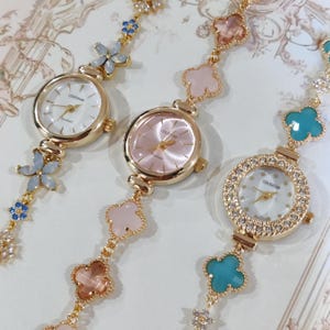 May include: Three gold wristwatches with different colored face and charm bracelets. The charms are four-leaf clovers in pink, blue, and turquoise. The watches have white, pink, and white faces with the word "COLISEUM" printed on them.