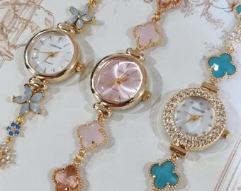 May include: Three gold wristwatches with different colored face and charm bracelets. The charms are four-leaf clovers in pink, blue, and turquoise. The watches have white, pink, and white faces with the word "COLISEUM" printed on them.