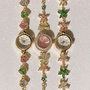 May include: Three gold-toned wristwatches with decorative bands. The watch faces are round with gold hands and the word "COLISEUM" printed on the face. The bands feature heart, flower, and bow-shaped embellishments in pink, green, and clear gemstones.