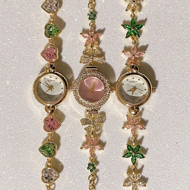 May include: Three gold-toned wristwatches with decorative bands. The watch faces are round with gold hands and the word "COLISEUM" printed on the face. The bands feature heart, flower, and bow-shaped embellishments in pink, green, and clear gemstones.