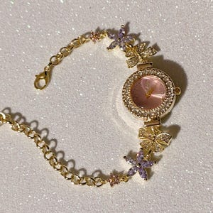 May include: Gold-toned wristwatch with a pink face and a bezel encrusted with clear crystals. The watch band features a delicate chain design, accented with flower and bow-shaped embellishments, adorned with crystals and purple and pink accents.