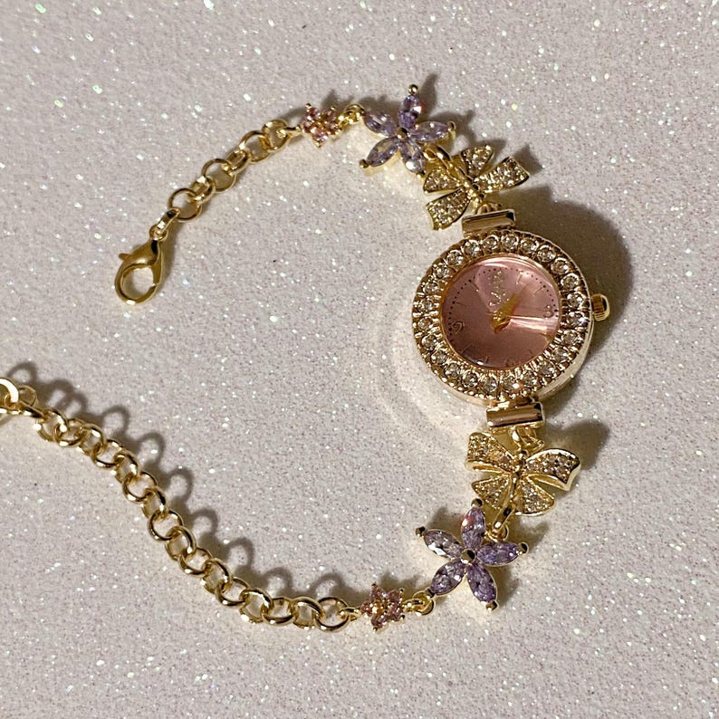 May include: Gold-toned wristwatch with a pink face and a bezel encrusted with clear crystals. The watch band features a delicate chain design, accented with flower and bow-shaped embellishments, adorned with crystals and purple and pink accents.