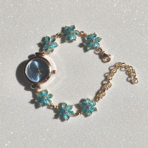May include: A gold-toned wristwatch with a light blue face and a floral bracelet. The bracelet features light blue flower-shaped accents. The watch has a delicate and feminine design, perfect for adding a touch of elegance to any outfit.