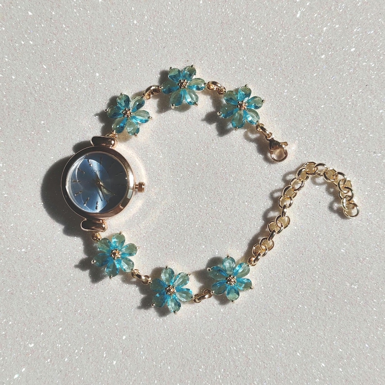 May include: A gold-toned wristwatch with a light blue face and a floral bracelet. The bracelet features light blue flower-shaped accents. The watch has a delicate and feminine design, perfect for adding a touch of elegance to any outfit.