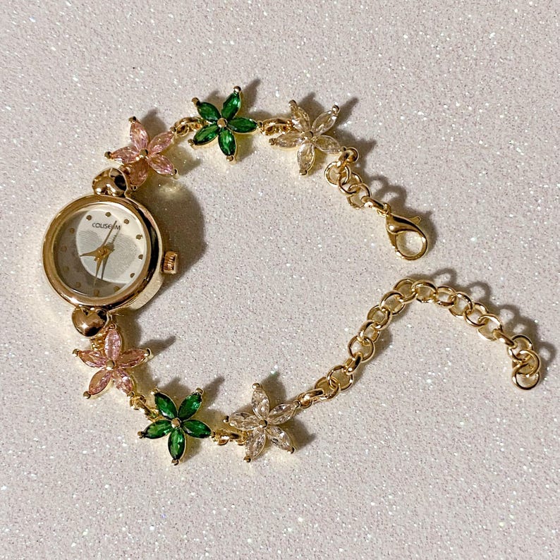 May include: Gold-toned wristwatch with a white face and delicate hands. The watch band is adorned with pink, green, and clear flower-shaped embellishments. The watch has a clasp closure and is set against a white, glittery background.