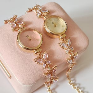 Puede incluir: Dos relojes de pulsera dorados con eslabones en forma de mariposa y detalles de cristal rosa y transparente. Las esferas de los relojes son redondas, con un dise&ntilde;o floral y una combinaci&oacute;n de colores rosa claro y dorado. Los relojes se muestran sobre una superficie rosa.