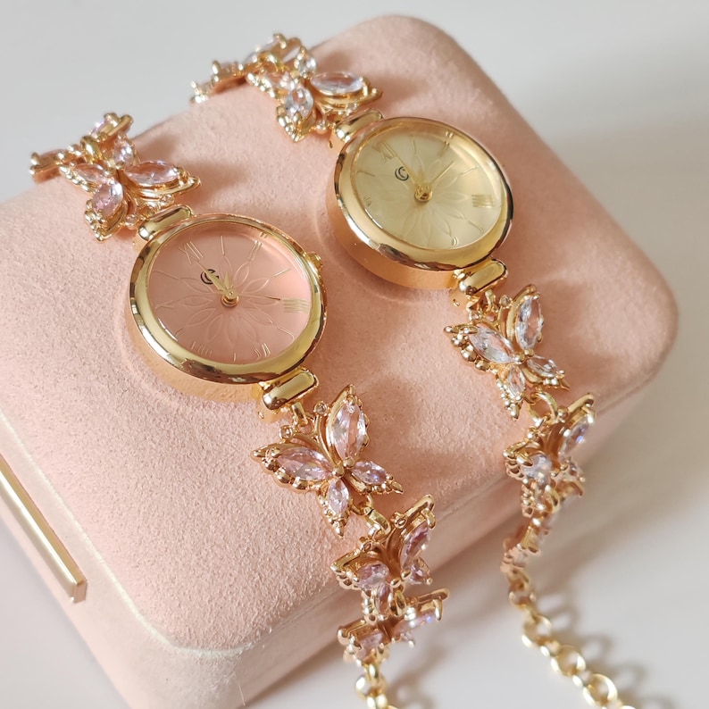 Puede incluir: Dos relojes de pulsera dorados con eslabones en forma de mariposa y detalles de cristal rosa y transparente. Las esferas de los relojes son redondas, con un dise&ntilde;o floral y una combinaci&oacute;n de colores rosa claro y dorado. Los relojes se muestran sobre una superficie rosa.