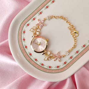 May include: A gold-tone watch with a pink face and a delicate chain bracelet adorned with pink flower accents.