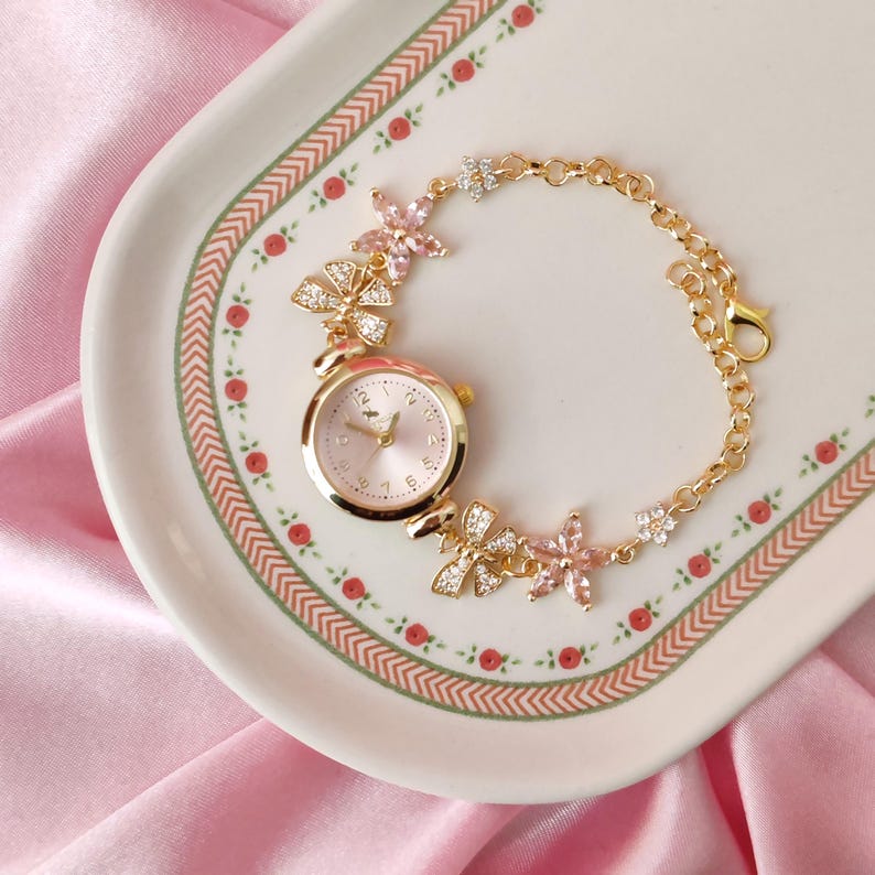 May include: A gold-tone watch with a pink face and a delicate chain bracelet adorned with pink flower accents.