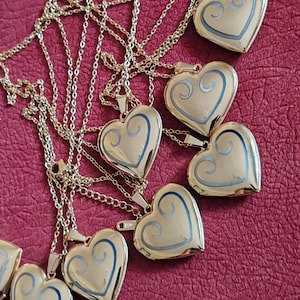 Puede incluir: Colgantes dorados en forma de corazón con delicadas cadenas. Cada colgante presenta un diseño azul en espiral. Los collares están dispuestos sobre una superficie roja, creando una exhibición visualmente atractiva de joyería.