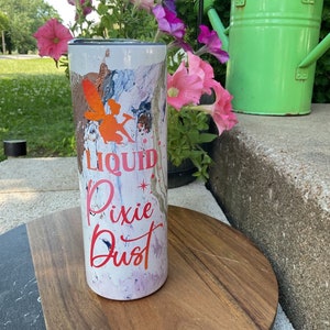 以下が含まれることがあります： カラフルな抽象的なデザインと「Liquid Pixie Dust」という赤い文字が書かれた白いタンブラー。タンブラーは黒い蓋と銀色の底をしています。