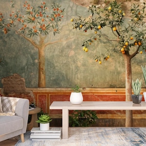 Op de afbeelding: Een muurschildering met een klassieke tuinscène met fruitbomen, waaronder sinaasappel- en citroenbomen. De muurschildering heeft een verweerde, verouderde uitstraling met een kleurenpalet van groen, oranje en bruin. De scène omvat een tafel, planten en een grijze fauteuil.