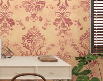 May include: A wall with a repeating burgundy damask pattern on a beige background. The pattern has a distressed, vintage look. A white desk with a wooden chair is in the foreground, with a plant and window blinds in the background.