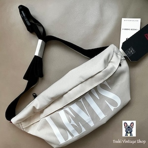 May include: Beige Levi's fanny pack with a black strap and the Levi's logo in white. The bag has a black tag attached. The bag is a neutral color and has a simple design.