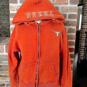 May include: Burnt orange zip-up hoodie with a hood and pockets. The word "TEXAS" is embroidered in white on the hood. A white Longhorn logo is on the chest. The hoodie has a full zipper and a ribbed hem and cuffs.