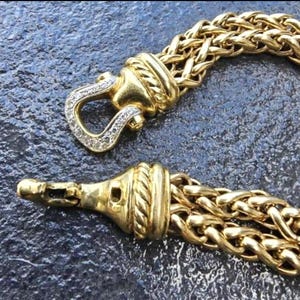 May include: Close-up of a gold-toned bracelet with a woven chain design. The clasp features a diamond-studded hook. The bracelet is displayed against a dark, textured surface, highlighting the jewelry's intricate details and luxurious appearance.