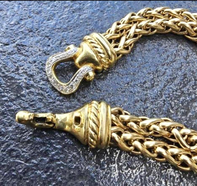 May include: Close-up of a gold-toned bracelet with a woven chain design. The clasp features a diamond-studded hook. The bracelet is displayed against a dark, textured surface, highlighting the jewelry's intricate details and luxurious appearance.