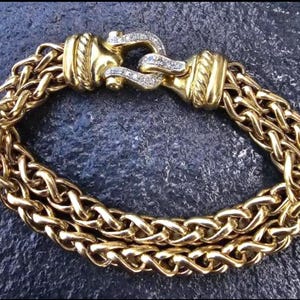 May include: A gold-toned bracelet with a woven chain design. The bracelet features a decorative clasp adorned with small, clear gemstones. The chain is made of multiple strands, creating a thick, textured appearance. The bracelet is a jewelry item.