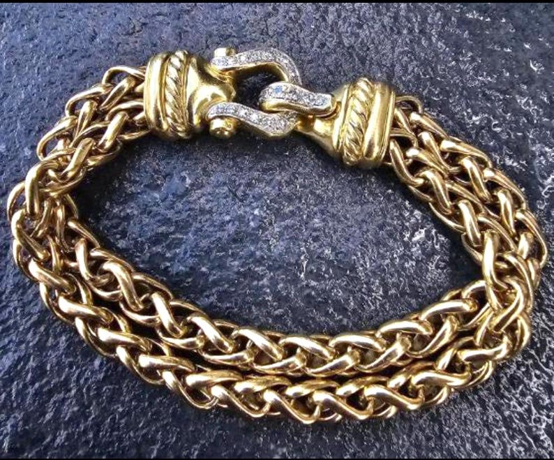 May include: A gold-toned bracelet with a woven chain design. The bracelet features a decorative clasp adorned with small, clear gemstones. The chain is made of multiple strands, creating a thick, textured appearance. The bracelet is a jewelry item.