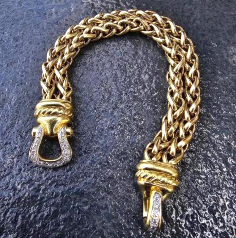 May include: A gold-toned bracelet with a woven chain design. The bracelet features a horseshoe-shaped clasp adorned with small, clear gemstones. The ends of the bracelet have decorative accents.