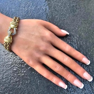 May include: Gold-toned bracelet with a braided chain design and a decorative clasp. The clasp features a horseshoe shape with embedded clear gemstones. The bracelet is displayed on a hand with manicured nails.
