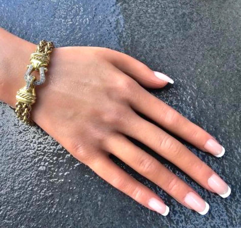 May include: Gold-toned bracelet with a braided chain design and a decorative clasp. The clasp features a horseshoe shape with embedded clear gemstones. The bracelet is displayed on a hand with manicured nails.