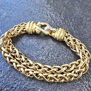 May include: A gold-colored bracelet with a woven chain design. The bracelet features a decorative clasp with small, clear stones. The bracelet is made of a shiny, metallic material.