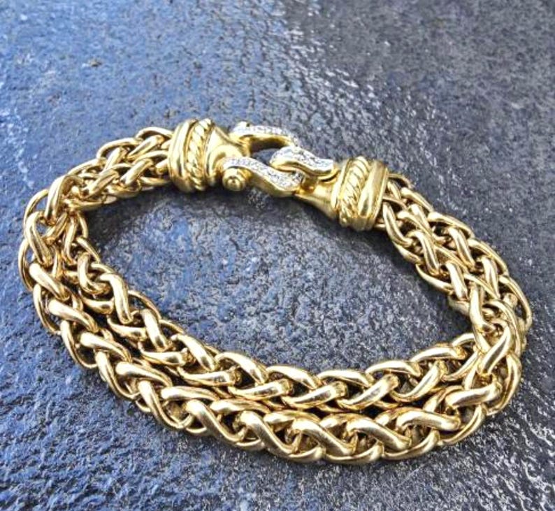 May include: A gold-colored bracelet with a woven chain design. The bracelet features a decorative clasp with small, clear stones. The bracelet is made of a shiny, metallic material.