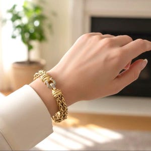 May include: A gold-toned bracelet with a braided design and a decorative clasp. The clasp features a horseshoe shape with small, clear stones. The bracelet is worn on a wrist, with a white shirt sleeve visible.